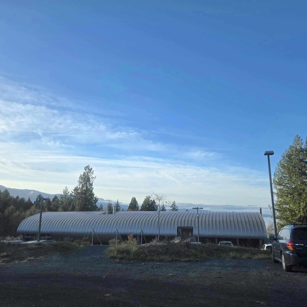 A morning shot of the VIU Deep Bay Marine Research Centre, which has a uniquely-shaped rounded, long metal roof. It looks to be the morning, and it is sunny out.