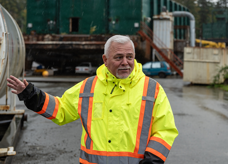 A man in high vis stands in front of a large derelict barge. He looks to his left with a slight smirk on his face. 