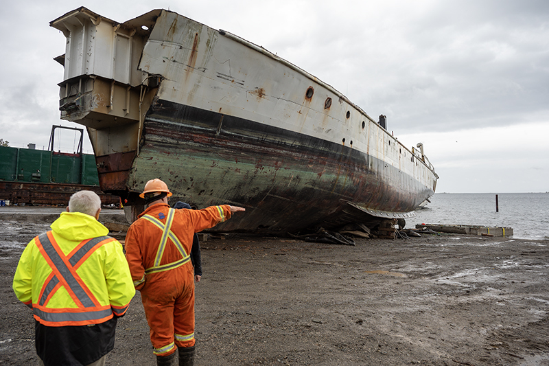 Two men in high visibility outfits stand facing away from the camera. One points at a large ship to his left.